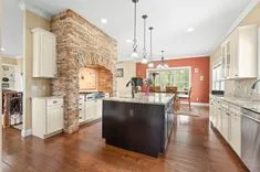 Spacious kitchen interior with stone archway over stove, hardwood floors, ivory cabinets, granite countertops, and a dining area with large windows.