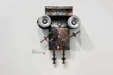 Wall-mounted assemblage resembling a robot face with round speakers for eyes, a rusty box for a head, and wires for limbs.