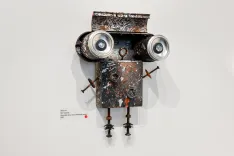 Wall-mounted assemblage sculpture resembling a robot face with circular eyes and rusted metal components.