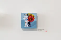 Figure of a unicorn with a colorful mane displayed on a canvas on a white wall, accompanied by a small red pushpin and artist attribution text.