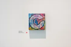 Colorful painting of a stylized "G" on a canvas displayed on a white gallery wall with descriptive text label to the side.