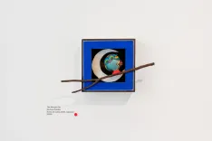 Modern art installation on a white wall featuring a blue square frame with concentric circles, a stylized globe in the center, and two brown rods crossing in front of the piece.