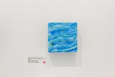 "Abstract blue ocean waves painting on a white gallery wall with a small informational plaque and red dot indicating the artwork is sold"