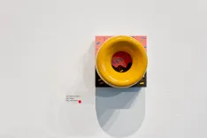 Modern art exhibit featuring a yellow funnel-shaped object on a colorful square panel mounted on a white wall with an informational label beside it.