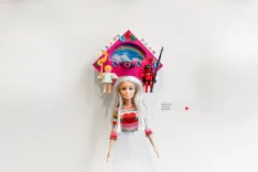 A display of various dolls mounted on a white wall, including one large doll in the foreground with a red and white outfit and two smaller dolls in a colorful house-shaped frame above.