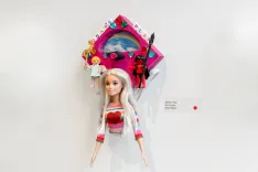 Contemporary art piece featuring a Barbie doll with mixed media elements including a miniature unicorn and other toys against a white gallery wall.