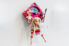 Barbie doll attached to a wall below a colorful dollhouse shelf with accessories.