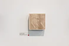 A minimalist art installation featuring a single, textured beige cube mounted on a white wall with a small red detail to the bottom left corner and a descriptive label at the bottom right.