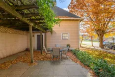 A cozy patio area with a metal table and chairs under a wooden pergola, next to a beige house with fallen autumn leaves on the ground and a vibrant orange tree beside the house during fall season.