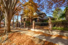 Autumn scene with a brown leaf-covered sidewalk leading to an elegant brick house with a large tree in the foreground and a wrought iron fence.