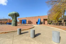 Outdoor public plaza with a large abstract green sculpture, cylindrical gray seats, a geometric blue sculpture by a brick building, and leafless trees against a clear blue sky.