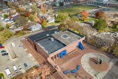 Aerial view of a modern brown building with distinctive blue art installations on the facade, surrounded by a parking lot, autumnal trees, and neighboring residential area.