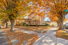 A sunny autumn day in a suburban neighborhood with a large house surrounded by trees with orange leaves, detached sidewalk, and a carpet of fallen leaves on the ground.