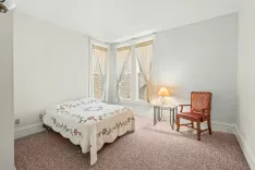Bright and simple bedroom with a double bed, patterned bedding, a side table with a lamp, a wooden armchair, light curtains, and beige carpet flooring.