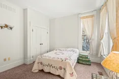 Bright and cozy bedroom with a single bed adorned with a floral quilt, a closet, a window with sheer curtains, vintage suitcases by the bed, and a lit table lamp beside a small mirror on the carpeted floor.