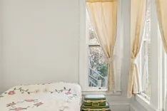 Bright bedroom with a white quilted bed, green suitcases, and cream-colored curtains framing an open window with a view of trees.
