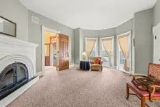 Spacious living room with plush carpet, a white fireplace, bay windows with curtains, classic wooden furniture, and a view into a hallway with a stained glass door.