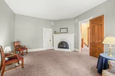 A spacious living room with a white fireplace, carpeted floors, and a mix of classic furniture including a rocking chair and a lamp on a table.