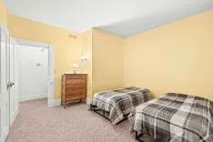 Brightly lit bedroom with yellow walls, two single beds with plaid bedding, a wooden dresser, and an en-suite bathroom entrance.