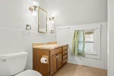Bright bathroom interior with white tile, a wooden vanity with a marble countertop, a toilet, and a window with a green curtain.
