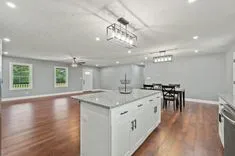 Spacious kitchen with white cabinetry, granite countertops, and modern lighting fixtures, adjacent to dining area with table set for four, all with hardwood flooring and a view of greenery through windows.