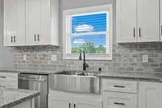 Modern kitchen interior with white cabinetry, stainless steel sink, grey subway tile backsplash, and a window view of blue sky and green trees.