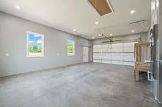 Empty garage interior with concrete floor, white doors, and windows showing outdoor greenery.