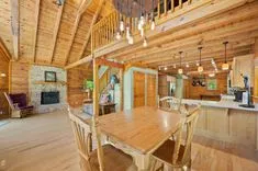 Spacious rustic log cabin interior with a dining table, kitchen bar area, and a stone fireplace.