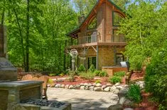 Two-story log cabin with stone foundation surrounded by lush green trees, featuring a second-story balcony, landscaped garden with stone path, and outdoor furniture.