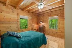 Cozy cabin bedroom with wooden walls, a ceiling fan, large bed with a teal bedspread, and a view of the forest through windows.