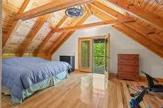A spacious attic bedroom with wooden beams and flooring, featuring a large bed, a ceiling fan, an exercise bike, a wooden chest of drawers, and French doors leading to a balcony.