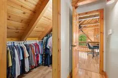 Interior view of a well-organized walk-in closet full of clothes with wooden finish, looking out through an open door towards a bright home office with a desk and chair.