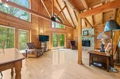 Interior of a spacious wooden cabin living room with exposed beams, large windows, and a stone fireplace.