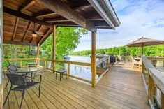 Spacious wooden deck of a lake house with outdoor furniture and a view of trees and a boat on the water.