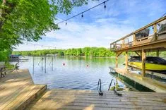 Sunny lakeside view with a wooden dock and a boathouse, string lights above, and lush greenery in the background.
