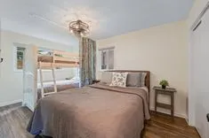 Brightly lit bedroom with a queen bed covered with a taupe comforter, a bunk bed in the background, wooden flooring, a nightstand with a lamp, and a ceiling fan.