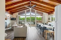 Bright modern living room with wooden beams, large windows, ceiling fan, and open-plan layout leading to a kitchen and dining area.