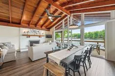 Modern open-air kitchen and dining area with wooden beams, a long dining table with chairs, and a view of lush greenery outside.