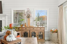 Cozy living room decorated with autumnal theme, featuring a wooden sideboard with seasonal decor, pumpkins, and a view of fall foliage outside the windows.