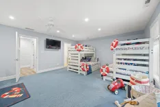 Spacious children's room with nautical theme, featuring bunk beds with lifebuoy decorations, a play area with toy car, and a flat-screen TV on the wall.