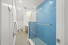 Modern bathroom interior with a blue tiled glass shower, white toilet, beige floor, wooden cabinet, and a framed mirror.