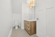 Modern bathroom interior with white walls featuring a toilet, wood-finished vanity with a sink, mirror, and wall sconce above.