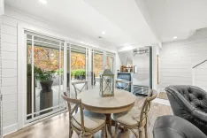 Bright, modern sunroom with sliding glass doors leading to a deck with autumn foliage, featuring a round wooden table, chairs, and a black leather sofa.