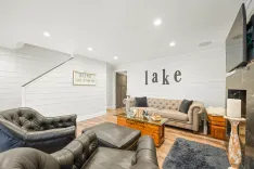 Modern cozy living room with neutral tones, shiplap walls with "lake" sign, tufted sofa, leather chairs, and a fireplace.