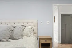A modern bedroom with a tufted headboard, neutral bedding, a wooden bedside table, and a closed gray door on a white shiplap wall.