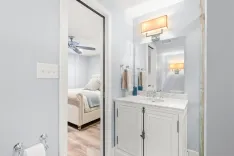 Interior view of a modern bathroom with white vanity and mirror leading to a bedroom with a ceiling fan and bed visible through the doorway.