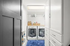 Modern laundry room interior with white cabinets, front-loading washer and dryer, with blue rug on floor and shelves with miscellaneous items above appliances.