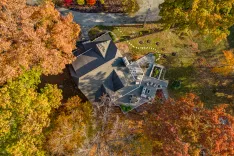 Aerial view of a large house with multiple gabled roofs surrounded by colorful autumn trees, featuring a backyard with a deck and outdoor furniture.