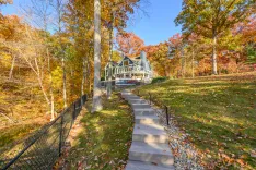 Alt: A large house with multiple gables surrounded by colorful autumn trees in a forest setting, with a wooden staircase leading up the hillside lawn to the back porch.