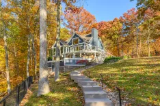 A large house with a wraparound deck surrounded by trees with autumn foliage, stone steps leading up a grassy hill.